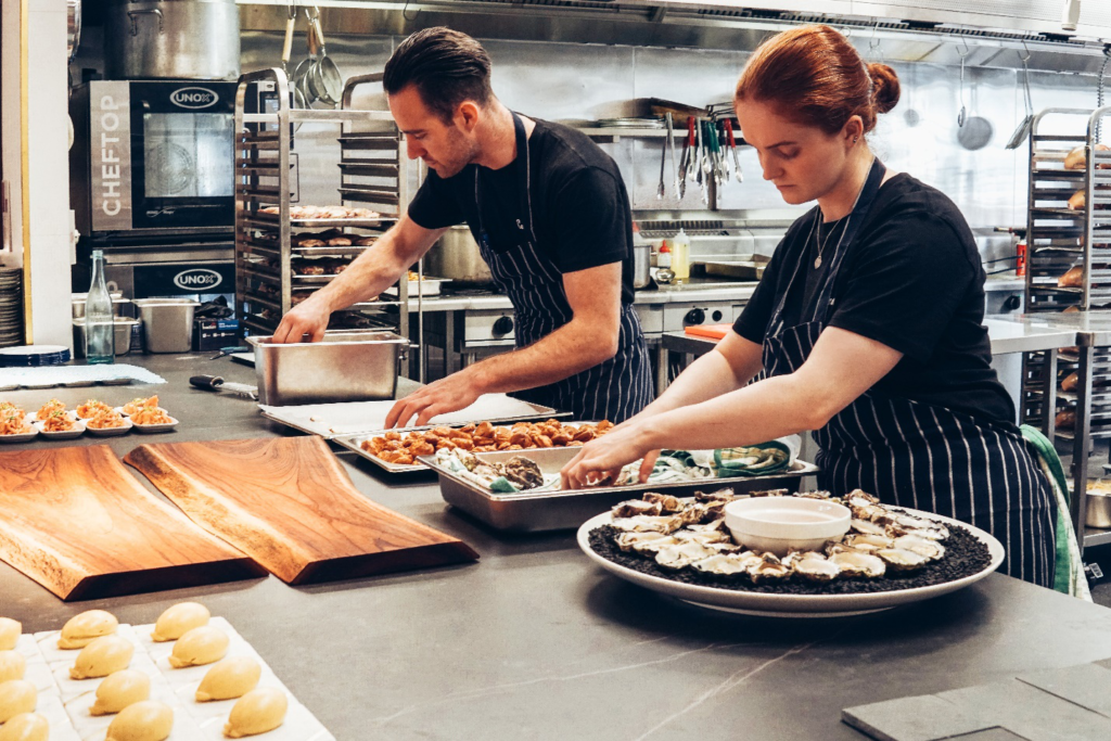 A man and woman stand together in a kitchen wearing black t-shirts and black and white striped aprons, preparing food and setting it up on platters in a unique catering service style.