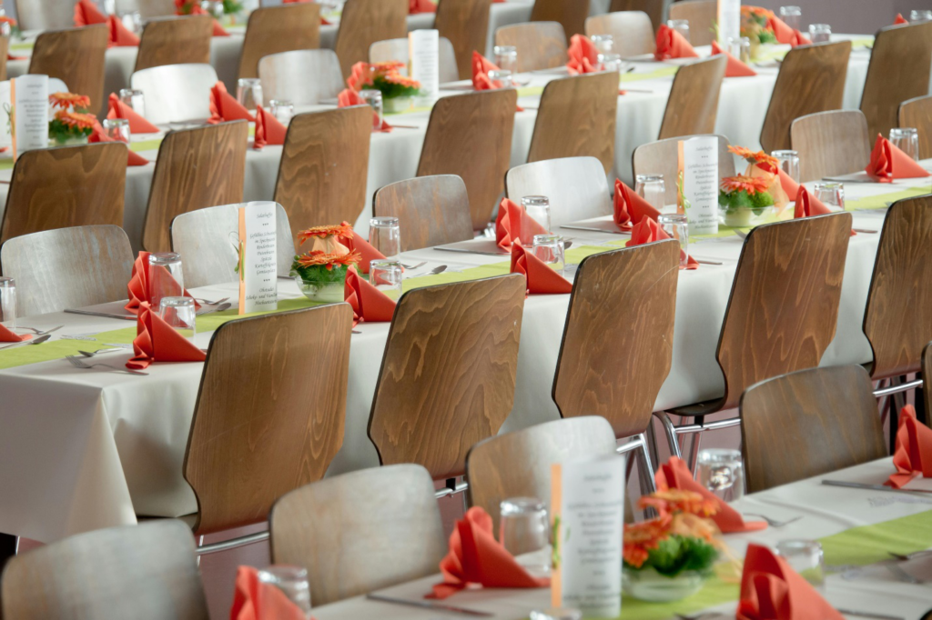 Brown chairs line tables covered with white tablecloths and decorated with orange napkins, silverware, and glasses, along with a standing menu, thanks to professional catering services.