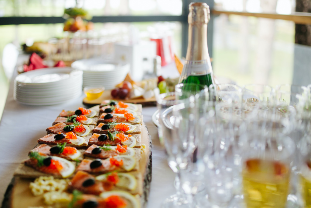 Customized starters on a wooden slab lay on a table next to champagne glasses and a bottle with plates and a cheeseboard showcase the importance of consulting a menu planning guide for catering.